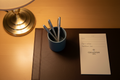 Overhead flat-lay of slate blue lacquer pen cup with hotel pens on leatherette desk pad with notepad and desk lamp shadow